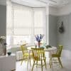 Floral-patterned roller blinds lowered over a three-pane bay window, filtering soft daylight; dining nook with round white table, four yellow chairs, pale grey-green walls and vase of purple flowers.