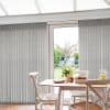 Grey textured vertical blinds cover floor-to-ceiling sliding glass doors, parted centrally to reveal a sunlit backyard; a small wooden dining table with chairs, flowers and pastries sits in the foreground.