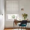 Beige linen Roman blind with subtle vertical stripes, lowered but slightly raised at the sill, filtering soft daylight into a compact study with desk, teal chair and vase of flowers.
