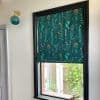 A floral-patterned teal Roman shade, lowered to cover the top half of a black-framed window and filtering light; nearby brass-and-glass wall sconce, pale hallway, and a small garden visible through the lower pane.