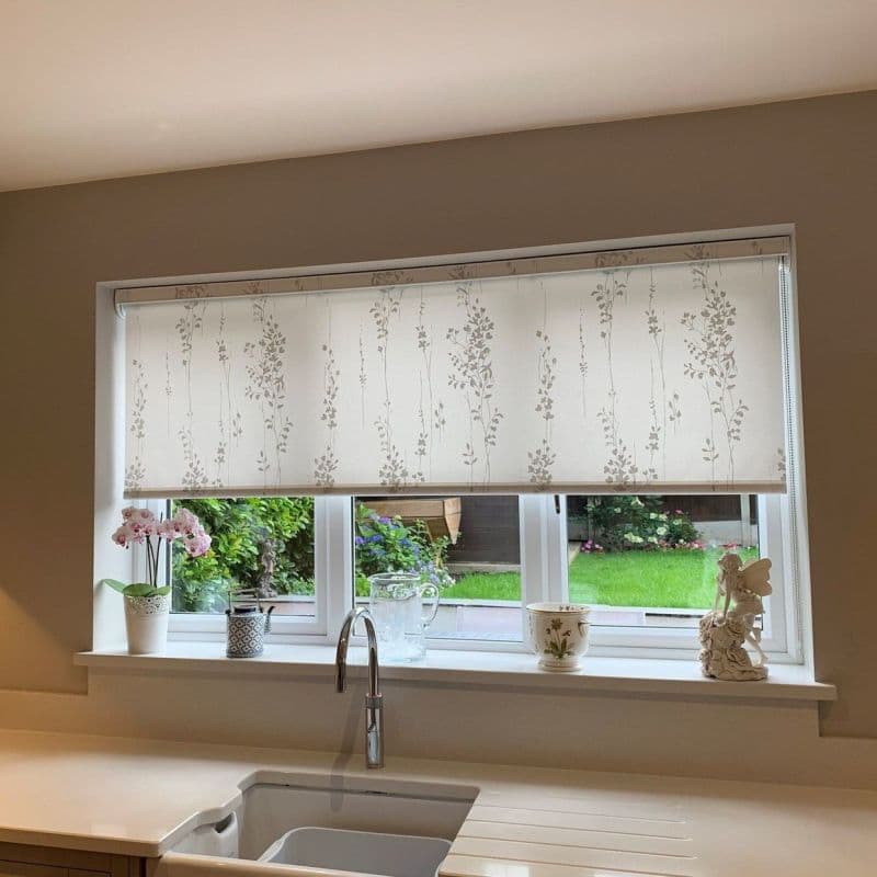 A patterned roller blind with vertical pale floral vines, lowered mid-window and filtering daylight; kitchen sink and counter below with potted plants, pitcher, and a green garden view.