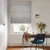 Grey textured Roman blind, lowered in soft folds over the window, filtering daylight; small desk with mirror, teal chair and vase of cascading flowers in a bright neutral home workspace.