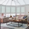 Light-blue cellular blinds cover tall conservatory windows, lowered and filtering soft daylight; a beige sofa, wooden coffee table and pastel cushions occupy a bright, cozy sunroom.