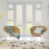Light beige pleated blinds cover French doors and sidelights, lowered to mid-height, filtering daylight and revealing lawn; bright sitting room with two rattan chairs, mint throw and geometric rug.