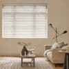 White horizontal Venetian blinds covering a large window, closed and softly diffusing daylight; living room with neutral sofa, wooden coffee table, vase, floor lamp, textured rug and warm ambient light.