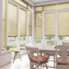 Beige horizontal venetian blinds covering tall corner windows, partly lowered and filtering soft daylight into a bright sunroom dining area with white table, chairs, floral cushions, and garden view.