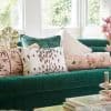 Emerald velvet sofa with assorted patterned throw pillows arranged along its backrest; bright bay windows flood the cozy living room with soft daylight and a leafy garden view.