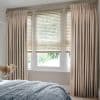 Beige patterned pinch-pleat curtains flank three lowered horizontal blinds whose angled slats filter soft daylight; small bedroom with a bed in the foreground and green foliage visible outside.