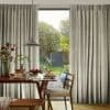 Pleated, floor-length grey curtains, drawn aside to frame a central opening and admit soft daylight; a wooden dining table set for breakfast beneath a pendant lamp, overlooking a sunlit garden.