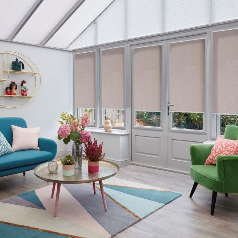 Beige fabric roller blinds lowered over tall glazed doors and windows, blocking most light but leaving lower panes exposed; bright conservatory-style living room with colorful sofas, rug, and garden view.