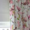 Floral fabric curtain — pleated and drawn at right with large pink and green blossoms — partially covering a lowered white roller blind, filtering soft daylight in a neutral-walled interior.