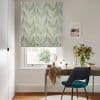 Roman fabric blind with green fern print lowered over a window, filtering daylight; desk with chair, vase of flowers and mirror beneath in a bright home office.