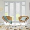 White internal Venetian blinds covering French doors and sidelights, partially lowered and filtering soft daylight; modern bright sitting area with two wicker chairs, pastel cushions, geometric rug and lawn view.