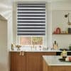 Striped dual-layer roller blinds lowered, alternating opaque and sheer bands; filtering daylight over a kitchen sink with brass faucet, white counters, wooden cabinets, and a brick-walled view outside.