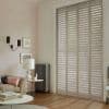 White full-height plantation shutters covering tall French doors, slatted louvers tilted to filter daylight; in a softly lit living room with pale walls, sofa, lamp, radiator, wooden floor.