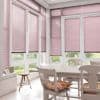 Pink horizontal Venetian blinds covering bay windows, lowered and filtering daylight (some panels slightly raised at bottoms), in a bright dining nook with white table and chairs and a garden view.