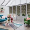 Decorative dark roller blinds with gold damask pattern, lowered over tall conservatory windows, partially blocking light; set in a bright sunroom with glass roof, colorful sofas, coffee table and flowers.