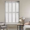 White louvered plantation shutters spanning a tall window, closed with slats slightly diffusing light; bedroom setting with upholstered headboard, bedside table with lamp and flowers, and an armchair.