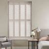 Cream-colored plantation shutters fill a tall window, fully closed with slats angled to filter soft daylight; set in a neutral bedroom with upholstered bed, round side table, lamp and armchair.