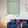 Light-green pleated cellular blind, fully lowered with horizontal folds; filtering daylight through a bottom gap, in a modern living room with a blue sofa, cushions and a floor lamp.