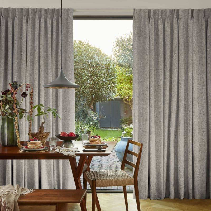 Grey floor-to-ceiling pleated curtains, parted at the center and hanging in soft folds, framing a glass door. Wooden dining table set for breakfast, pendant lamp, sunlit garden beyond.