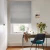 Grey fabric Roman blind lowered to mid-height, folded and filtering daylight; bright home office with a wooden desk, teal chair, tabletop mirror, vase of flowers and framed seaside print.