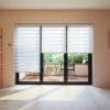 Three striped dual‑layer roller blinds over sliding glass doors, alternating sheer and opaque bands; partially lowered, filtering daylight onto pale wood‑floored room overlooking small balcony with table, chairs and greenery.
