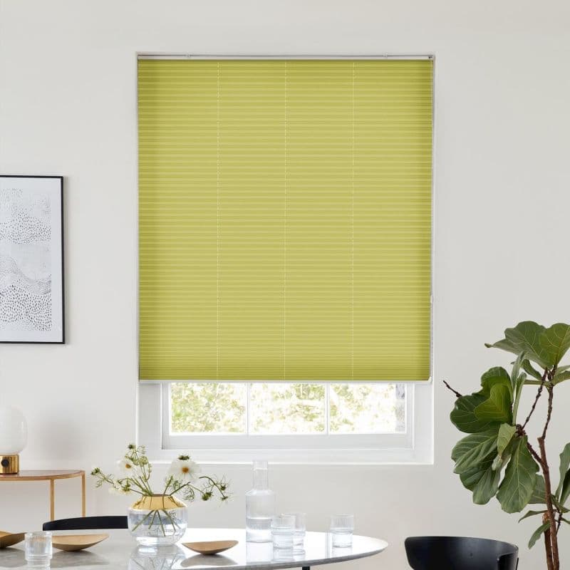 A lime-green pleated blind, fully lowered across a recessed window with vertical seams, blocking most light; modern dining area with white walls, table with glassware, flowers and a potted plant.