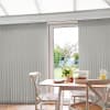 Grey fabric vertical blinds, floor-to-ceiling with slim slats and visible headrail; mostly closed but parted centrally, filtering daylight into a bright dining nook with wooden table, chairs, and a garden view.