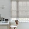 Beige semi-sheer Roman shade lowered across a multi‑pane window, horizontal folds visible; filtering diffuse daylight; minimal home office with white desk, wooden chair, pendant lamp and faint tree silhouettes outside.