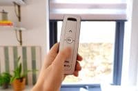 A zebra-style roller blind, partially lowered with alternating opaque and sheer stripes, filtering daylight. A hand holds a remote toward a bright room window with plants and shelves.
Text transcribed: ↑ my omfy