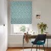 Teal-and-white diamond-patterned Roman blind lowered over a window, filtering daylight; in a small home office, a desk with mirror, vase of flowers, scarf-draped surface and teal-upholstered chair.