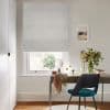 Light-grey, vertically ribbed Roman blind lowered over the window, filtering soft daylight; simple home office with wooden desk, teal chair, vase of flowers and framed seaside print.