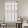 White plantation shutters spanning a tall recessed window, divided into four panels with horizontal louvers, closed and diffusing daylight; neutral bedroom setting with upholstered armchair, bedside table and lamp.
