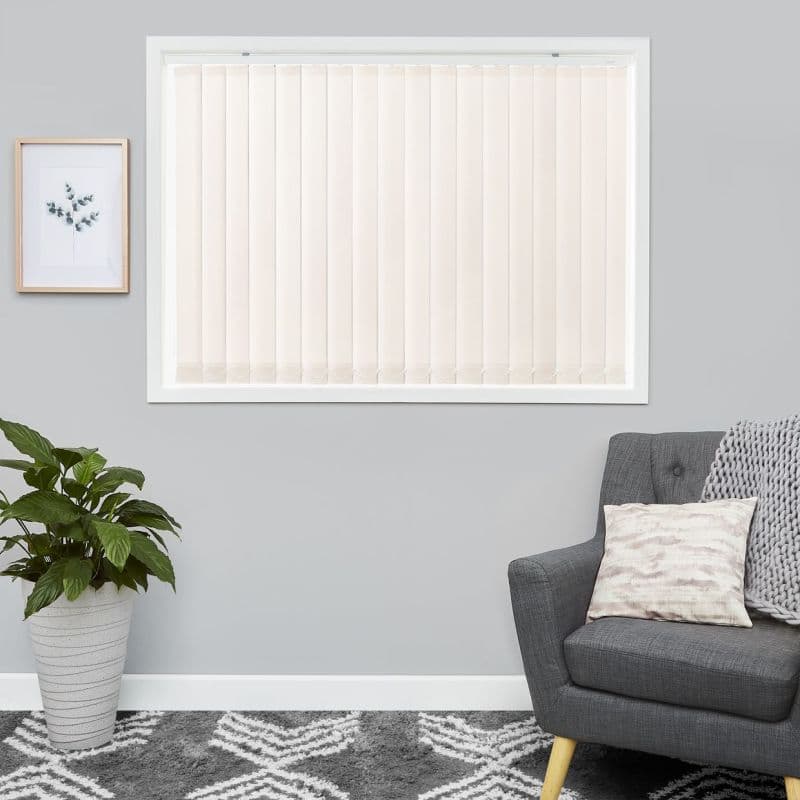Beige vertical blinds fill a centered rectangular window—evenly spaced fabric slats with bottom chains—closed and softly filtering daylight; grey living room with framed botanical print, potted plant, armchair, patterned rug.