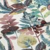 Botanical-print fabric with a white background and multicolored leaves (teal, olive, mauve), folded so an edge overlaps the center; close-up studio view with soft, even lighting.