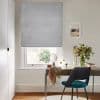 Light-grey fabric Roman blind, fully lowered, filtering soft daylight through a narrow gap; above a wooden desk in a bright office/vanity with a teal chair and a vase of flowers.