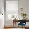 White Roman blind, lowered and slightly folded, filtering soft daylight; above a desk with teal chair, mirror and vase of flowers in a neutral-walled room, window view of greenery.