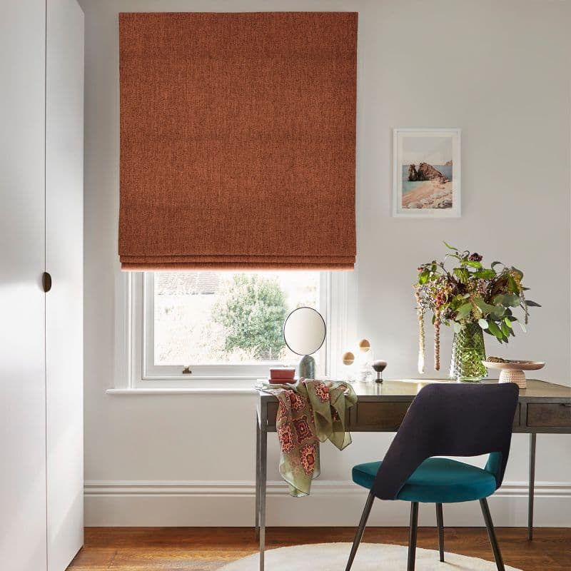 A rust-colored fabric Roman blind lowered over a window, slightly folded at the bottom, filtering daylight into a bright, neutral home office with a desk, teal chair, and a vase of flowers.
