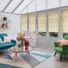 Patterned roller blinds (cream damask) cover tall rectangular windows and glass door, mostly lowered and filtering daylight; in a bright conservatory-style living room with teal sofa, green armchair, floral centerpiece.