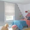 Star-patterned fabric roman blind fully lowered over the window, white with small multicolored stars, filtering soft daylight; in a girl's bedroom with gray/pink walls, blue quilt, string lights, wicker toy and pom‑pom pouf. LOVE