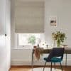 A beige textured Roman blind lowered over a sash window, gently filtering daylight; above a wooden desk with teal chair, vase of flowers and framed seaside print in an office.