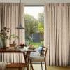Patterned, pleated full-length curtains, pulled slightly open to reveal a central glass door; hanging and filtering soft daylight. Wooden dining table set for breakfast with view of a green garden beyond.