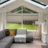 Pleated cellular blinds across a triangular-topped bay window, partially lowered to mid-height and filtering daylight; set in a bright conservatory-style sitting room with grey L-shaped sofa, floor lamp, and lawn view.