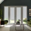 French doors with star-patterned roller blinds block sunlight as a glass coffee table and chevron rug sit between two green armchairs in a softly lit living room with plants.