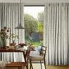 Floor-length pleated curtains with pale leafy print, drawn aside at center to reveal a sunlit garden; dining table set for breakfast beneath a hanging pendant lamp.