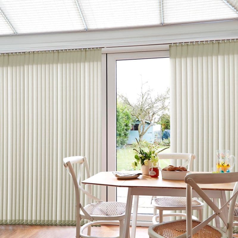 Cream vertical blinds covering sliding glass doors, floor-to-ceiling with evenly spaced slats, mostly closed but parted at center; sunlit breakfast/dining nook with wooden table, woven-seat chairs, garden view.