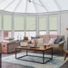 Pale green pleated cellular blinds, lowered and closed, filtering soft daylight across tall conservatory windows; cozy living room with beige sofa, chunky knitted throw, wooden coffee table, and blue rug.