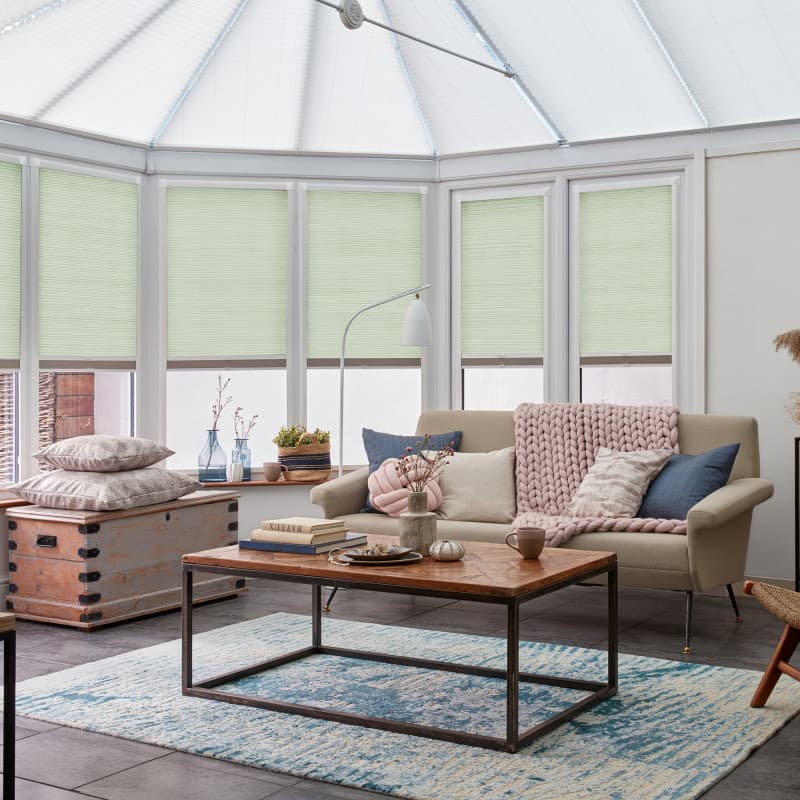 Pale green pleated cellular blinds, lowered and closed, filtering soft daylight across tall conservatory windows; cozy living room with beige sofa, chunky knitted throw, wooden coffee table, and blue rug.