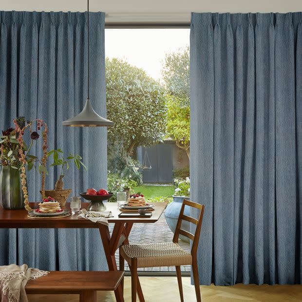 Blue pleated full-length curtains, hung and drawn slightly apart to reveal a glass patio door, framing a sunlit backyard; dining table with pendant lamp, pancakes and wooden chairs in foreground.